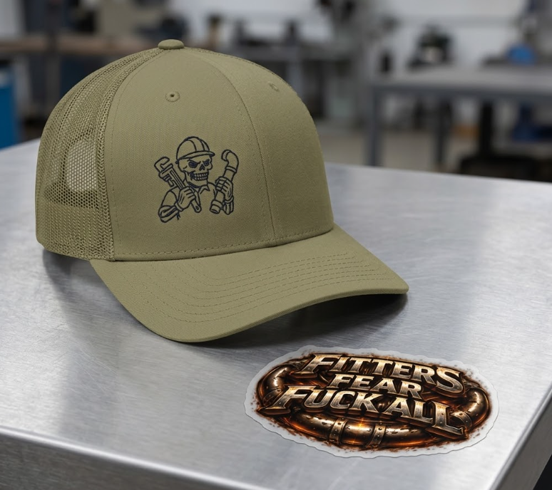 An olive green trucker hat embroidered with a skeleton plumber graphic sits on a stainless steel surface next to an industrial-style sticker featuring the text "FITTERS FEAR FUCK ALL" over bronze pipes.